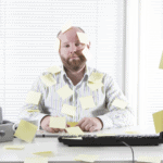 man is sitting at desk with a computer on it looking stuck with post it notes wondering "where is your schedule"